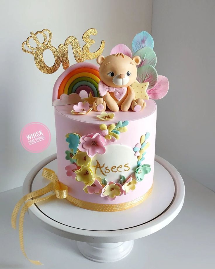 a pink cake with a teddy bear and rainbow decoration on top is sitting on a white pedestal