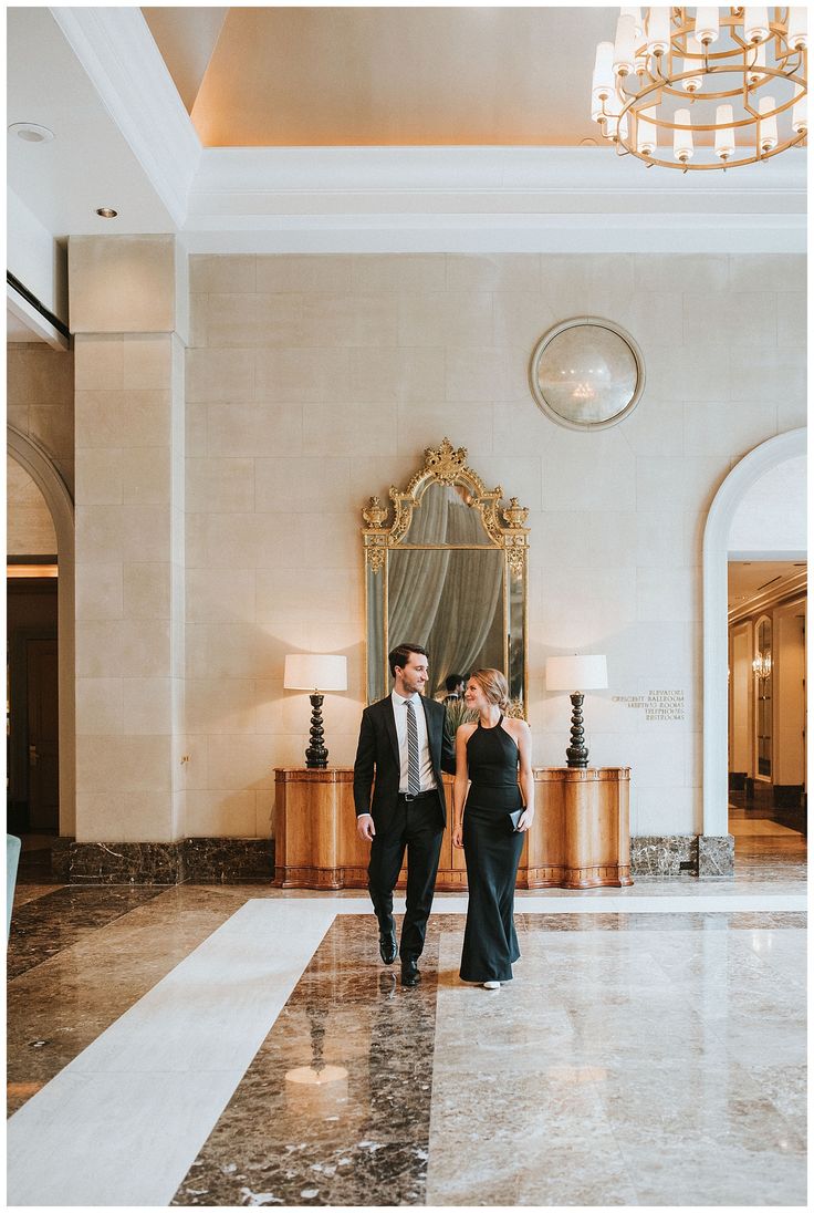 a man and woman in formal wear walk through the lobby of a hotel with chandelier