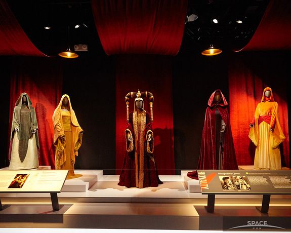 several costumes are on display in a museum case with red drapes and curtains behind them