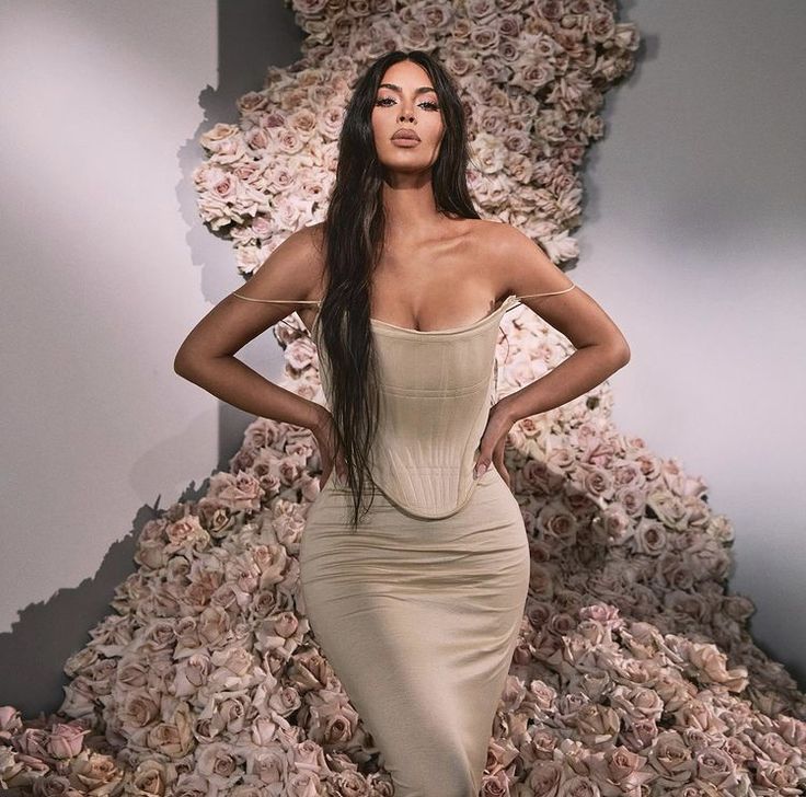 a woman standing in front of a pile of flowers wearing a tan dress with her hands on her hips