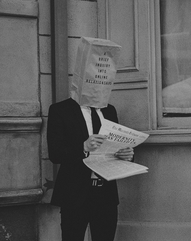 a person with a paper bag on their head standing in front of a building reading a newspaper