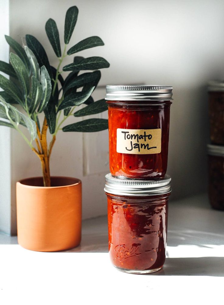 two jars of jam sitting next to a potted plant on a table with the label tomato jam written on it