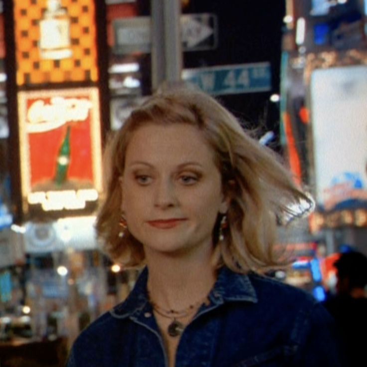 a woman with blonde hair wearing a denim shirt and earrings standing in front of neon signs