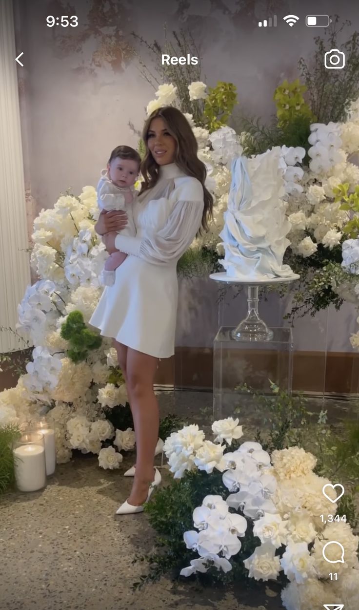 a woman holding a baby standing in front of flowers