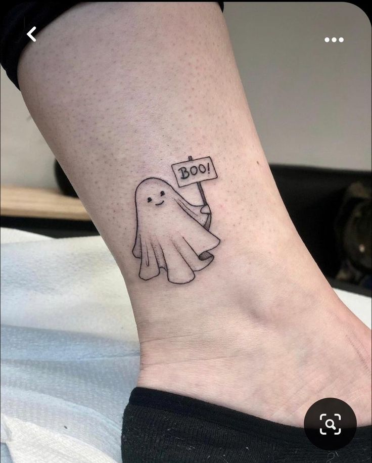 a small black and white ghost holding a boo sign tattoo on the ankle by kristien voddon