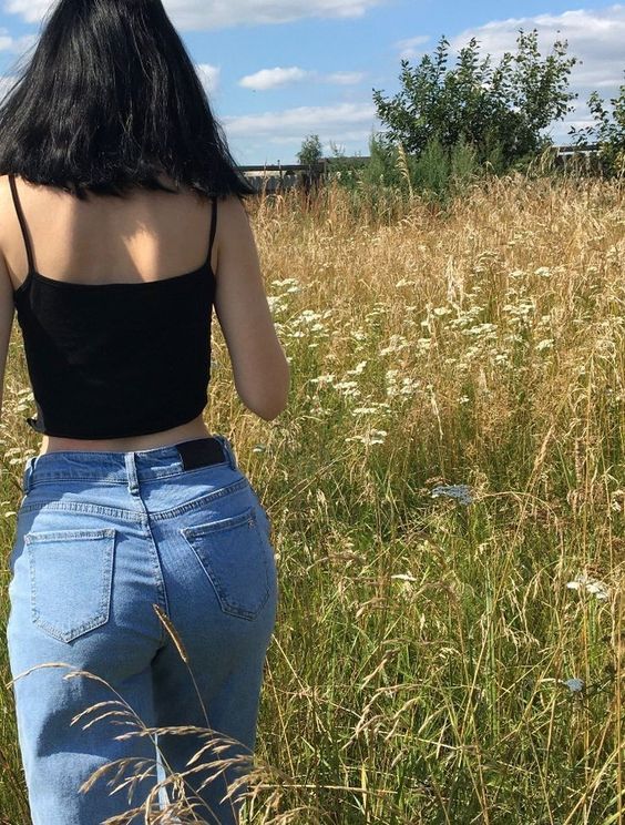 a woman standing in tall grass with her back to the camera, wearing jeans and a black tank top