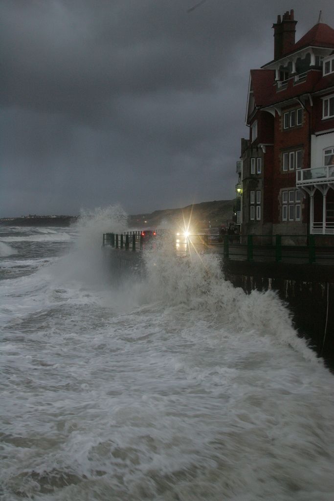 the waves are crashing into the shore as cars drive by on the road in front of houses