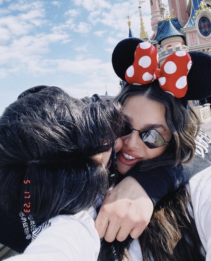 two people wearing minnie mouse ears hugging each other in front of a castle with a sky background