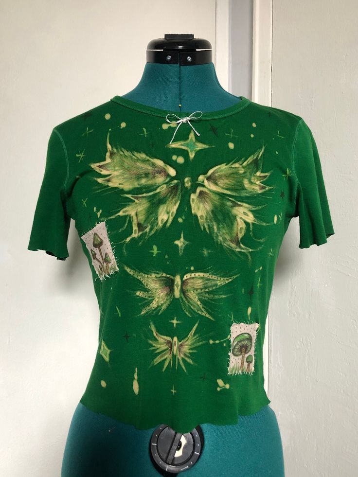 Bleach-painted kelly green T-shirt with butterflies, mushrooms, and stars graphic. Patches are adhered with fabric glue and sewn on. Small bow detail on the collar. All graphic details are done in fabric marker. Should fit sizes XS-S. This piece is handmade, handpainted, one of a kind, and made from a reused T-shirt. Wash inside out or carefully handwash in cold water.  In case of accidents, free repair is offered. Price is calculated based on hours spent making the product (minimum wage), effor Green Graphic Print Y2k Tops, Y2k Green Graphic Print Tops, Green Y2k Graphic Print Tops, Fitted Fairy Grunge T-shirt For Spring, Fairy Grunge Fitted Short Sleeve T-shirt, Fitted Green Y2k T-shirt, Spring Fairy Grunge Fitted T-shirt, Fairy Grunge Fitted T-shirt For Spring, Fitted Crew Neck Fairy Grunge T-shirt