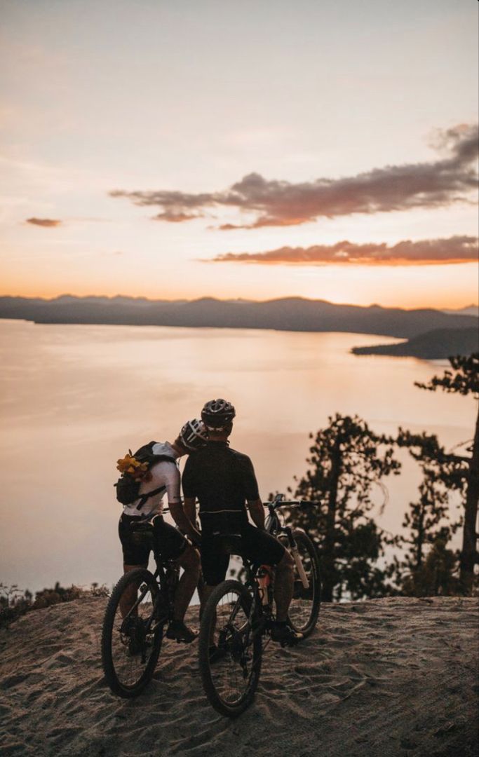 two bicyclists on top of a hill overlooking the water at sunset with trees in the foreground