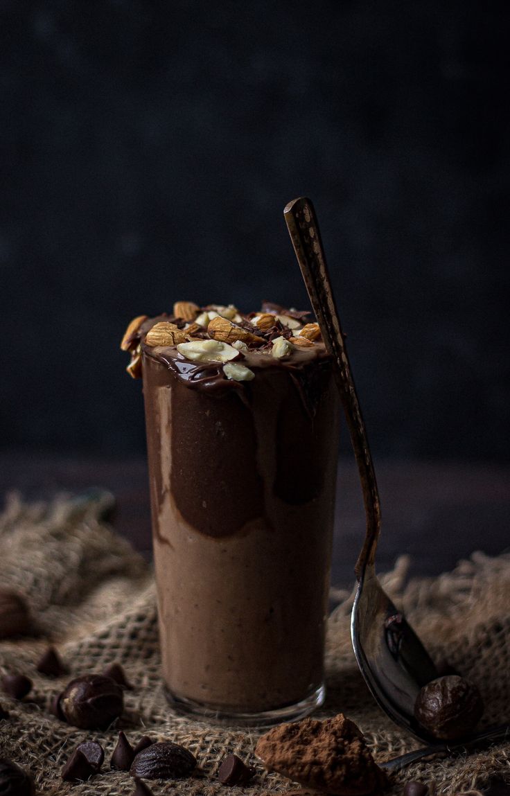 a chocolate dessert in a glass with a spoon on the side and nuts scattered around it