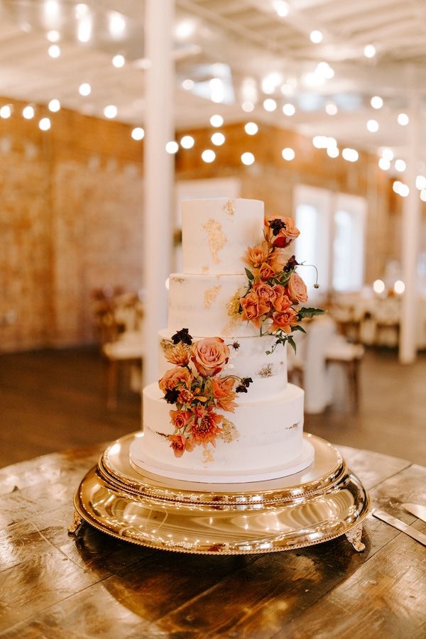 a white wedding cake with orange flowers on top sits on a gold platter in the middle of a banquet hall