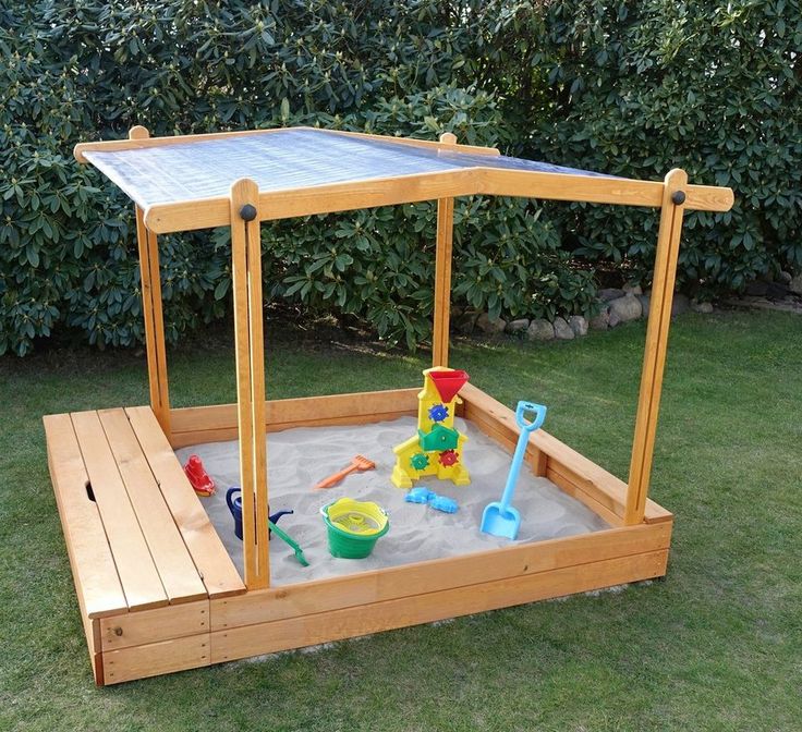 a wooden sandbox with toys in it on the grass next to some bushes and trees