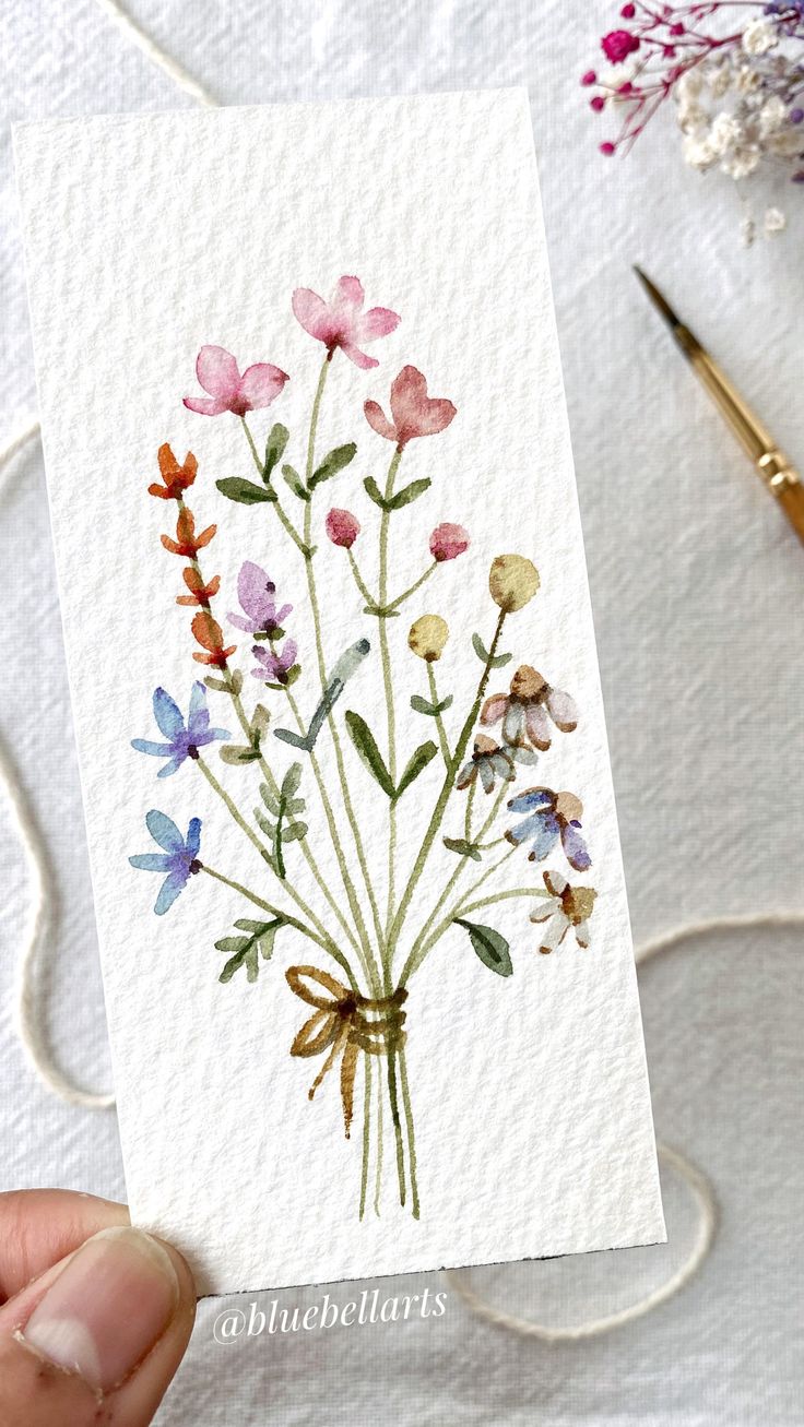 a person holding up a card with watercolor flowers on it next to some scissors