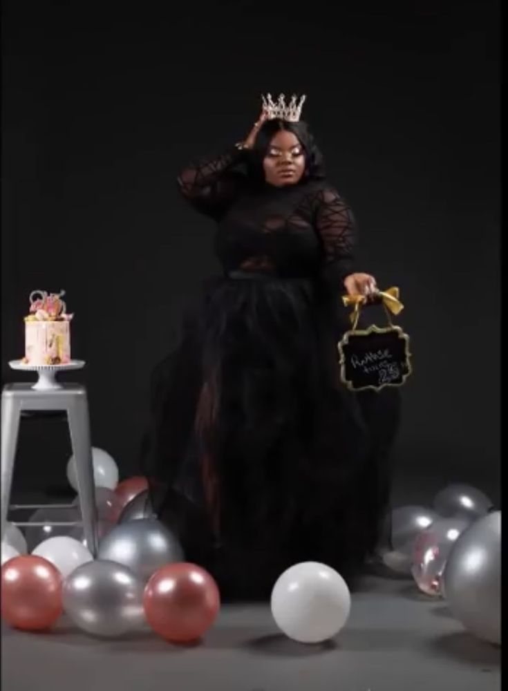 a woman in a black dress standing next to balloons and cake with a crown on her head