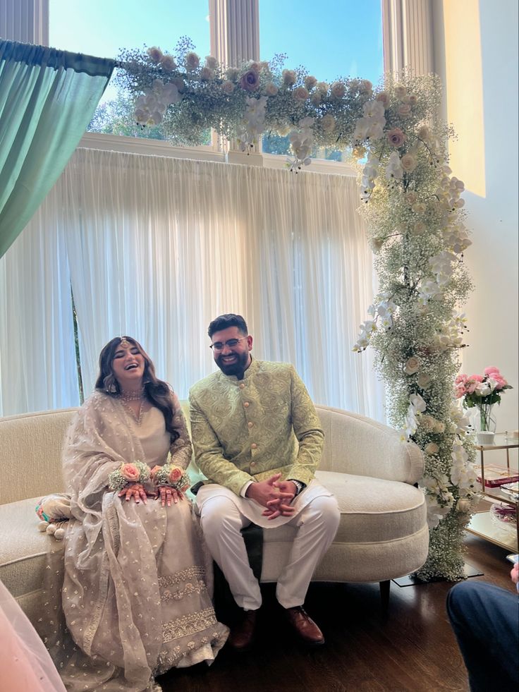 a man and woman sitting on a couch in front of a window with floral decorations