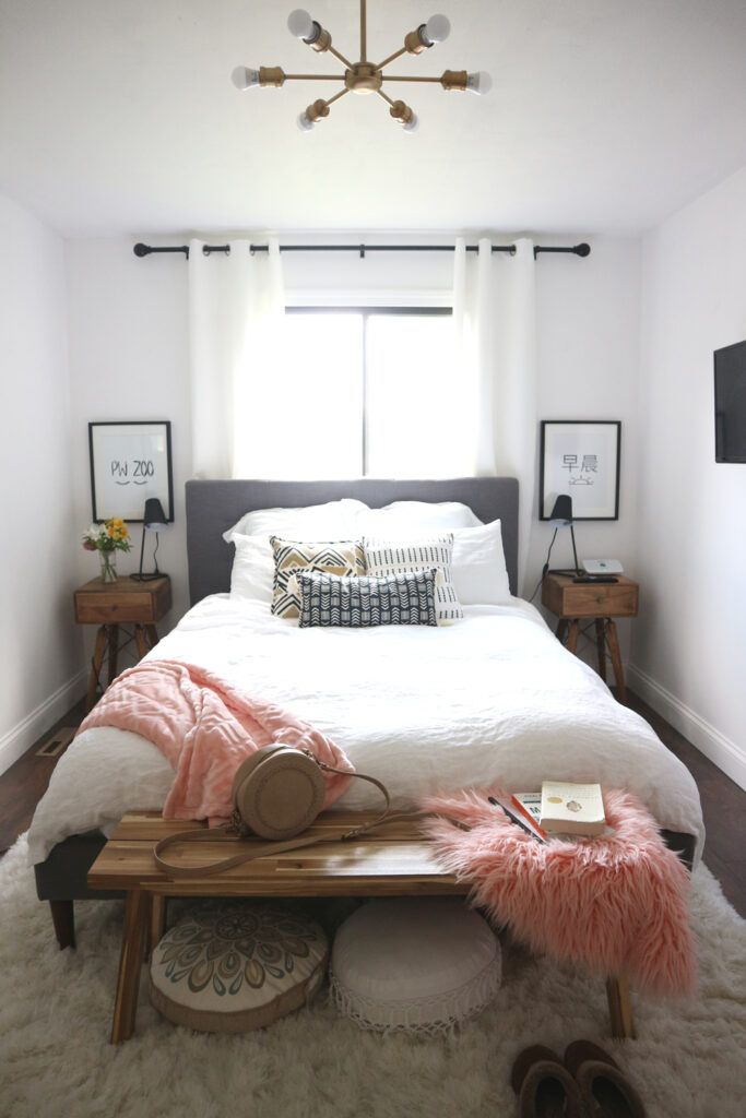 a white bed sitting under a window next to a wooden table with a lamp on top of it