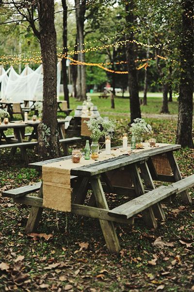 an outdoor picnic table set up with mason jars and greenery on it, surrounded by trees