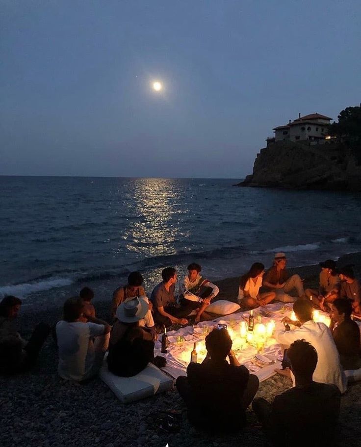 a group of people sitting around a table on the beach at night with a full moon in the background