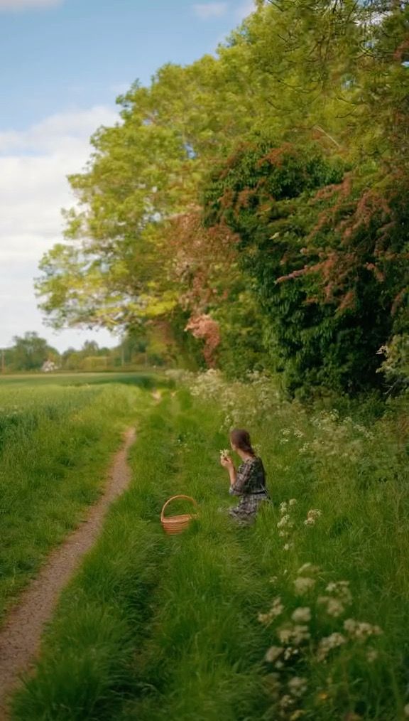 a woman sitting on the side of a dirt road next to a lush green field