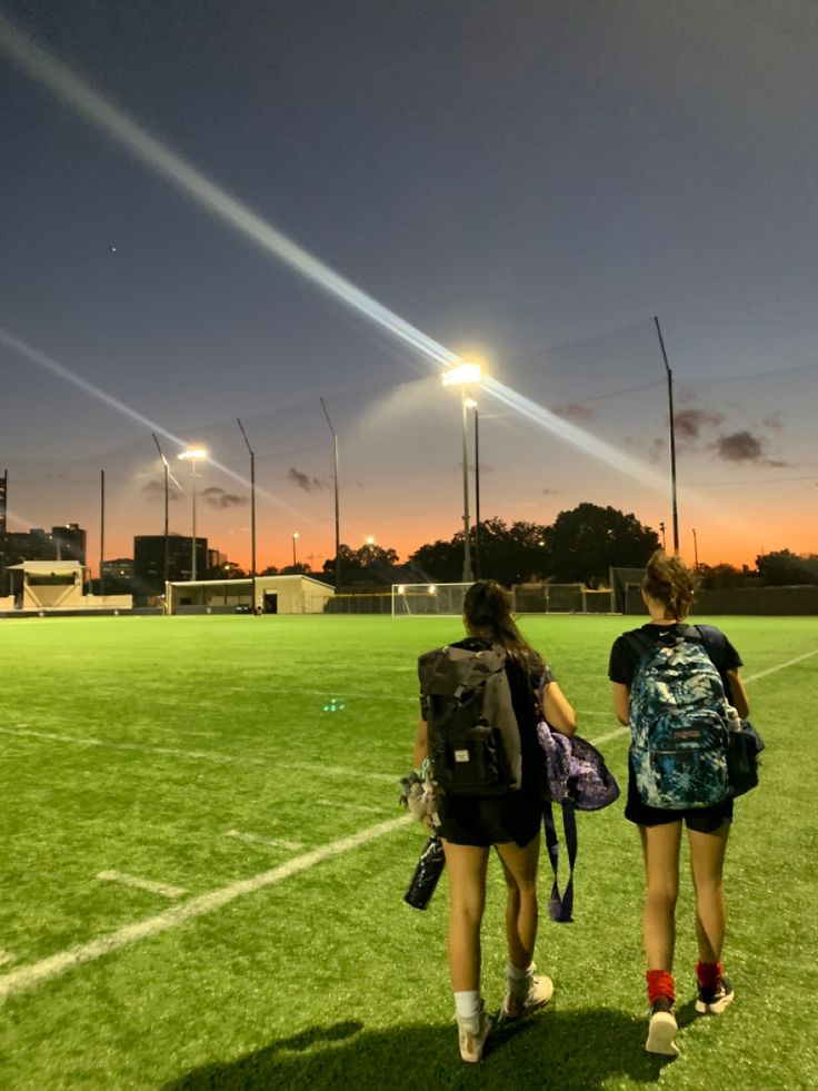 two girls walking across a soccer field at night with their backpacks on and one carrying a book bag