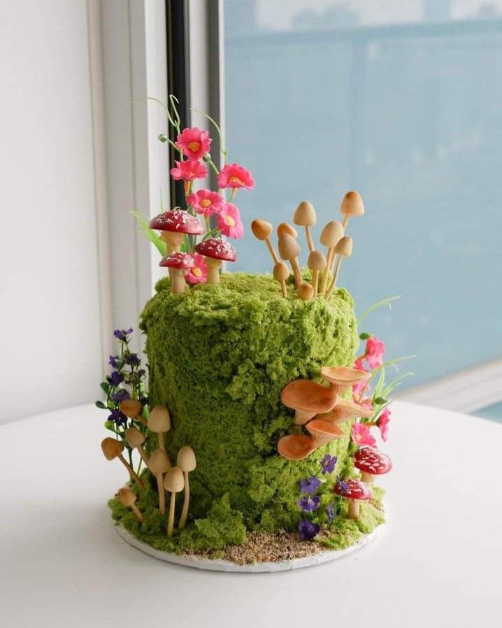 a green cake with mushrooms and flowers on it sitting on a table next to a window