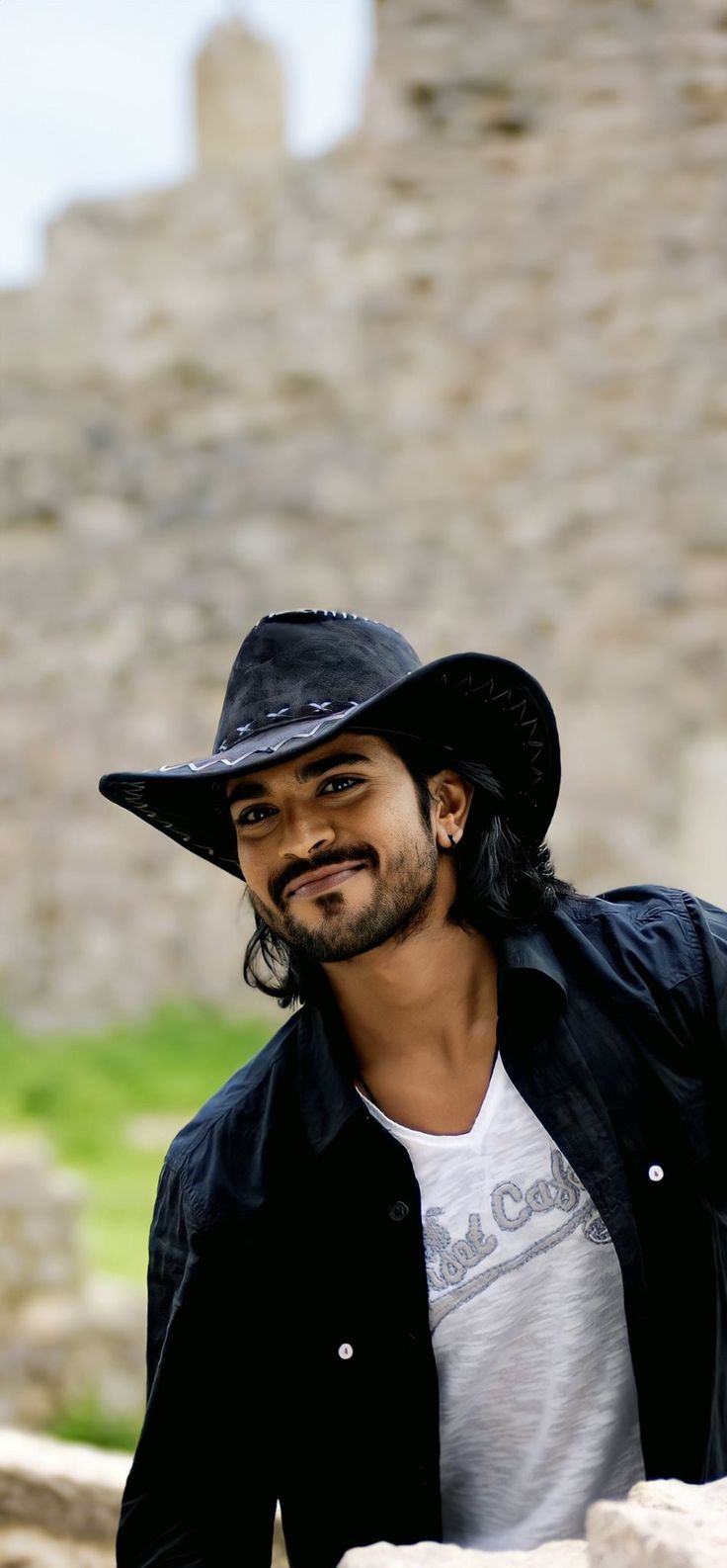 a man wearing a black hat standing next to a stone wall with a castle in the background