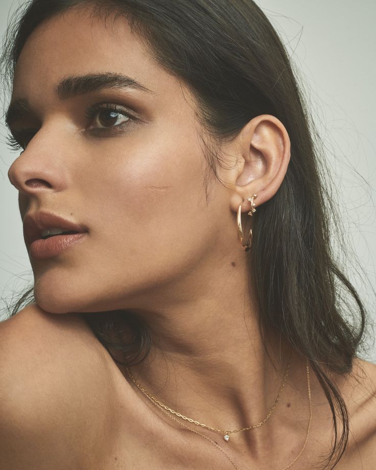 a close up of a woman wearing gold earring and necklace with her right hand on her shoulder