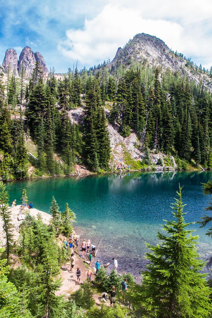 a group of people standing on the shore of a lake surrounded by trees and mountains