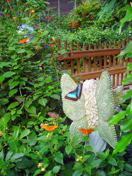 a butterfly statue sitting on top of a wooden bench in the middle of flowers and plants