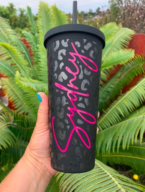 a person holding up a black cup with pink writing on it and palm trees in the background