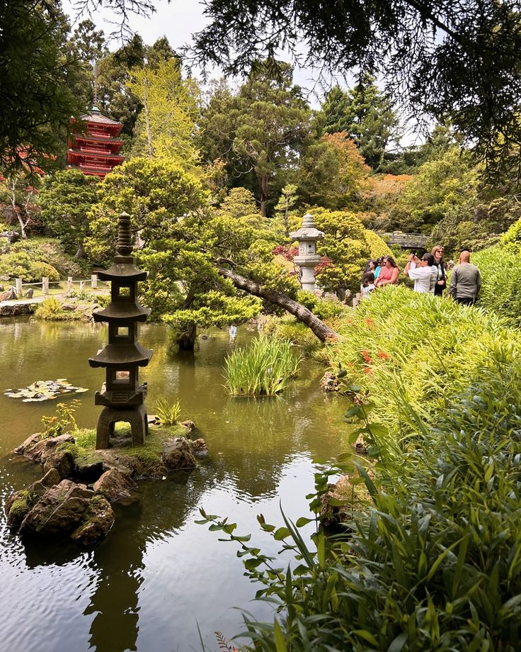 a small pond surrounded by trees and rocks in a garden with people walking around it