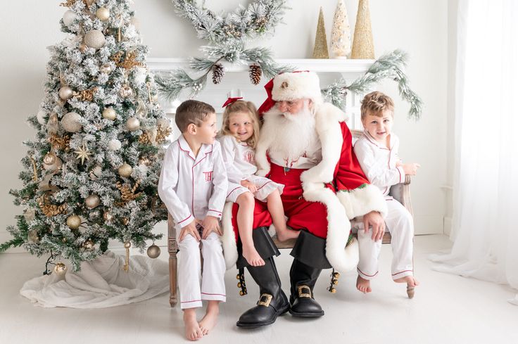 a santa clause sitting on top of a chair next to two small children in front of a christmas tree