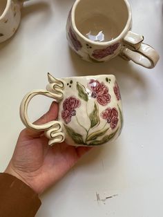 a hand holding a coffee cup with handles and flowers painted on it, next to two mugs