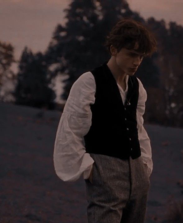 a young man standing on top of a grass covered field next to a tree filled hillside