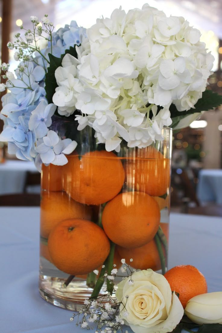 an arrangement of flowers and oranges in a glass vase on a white table cloth