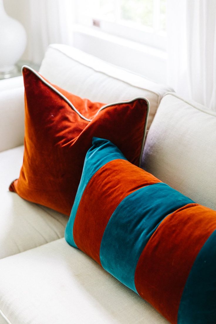 two orange and blue pillows sitting on top of a white couch next to a window