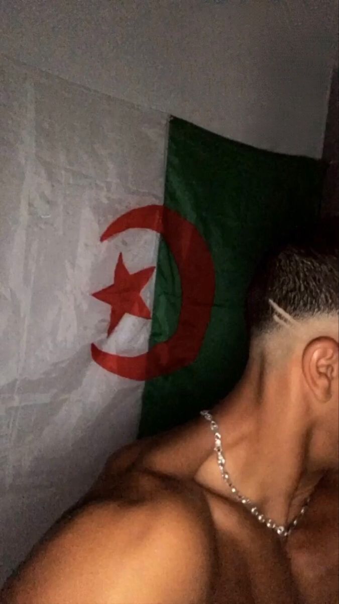 a man with no shirt on laying in bed next to a flag and a chain around his neck