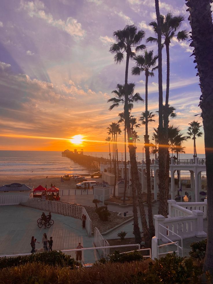 the sun is setting at the beach with palm trees and people walking on the boardwalk