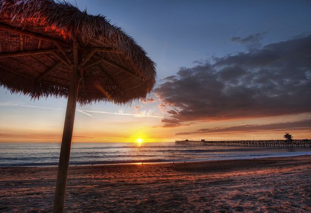 the sun is setting at the beach with palm umbrellas on the sand and water in the background