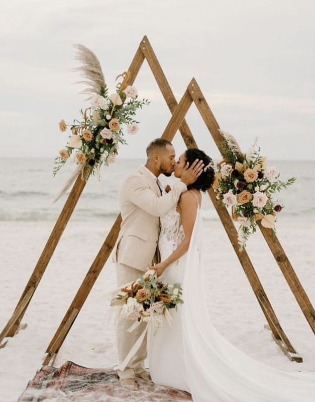 a bride and groom kissing in front of an arch made out of wooden sticks on the beach