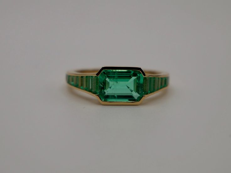 At the centerpiece of this lab grown Colombian emerald ring is a radiant 1.5 ct emerald, set east west to highlight its natural allure. Flanking it are calibre cut emeralds, each uniquely shaped to create a seamless gradient effect, making every ring truly one-of-a-kind. Set in luxurious 18K yellow gold, this ring exudes timeless charm. What makes it truly special? Calibre cut stones are individually shaped to fit precisely into the design, resulting in a mesmerizing display of color and brillia Timeless Green Emerald Ring With Center Stone, Heirloom Green Baguette Cut Emerald Ring, Timeless Rectangular Prong Set Emerald Ring, Timeless Rectangular Emerald Ring With Prong Setting, Gia Certified Green Square Cut Jewelry, Gia Certified Green Emerald Art Deco Ring, Modern Emerald Rings With Baguette Cut, Luxury Square Cut Green Emerald Ring, Classic Green Radiant Cut Ring