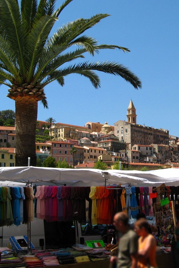 people walking around an open air market with palm trees in the foreground and buildings in the background