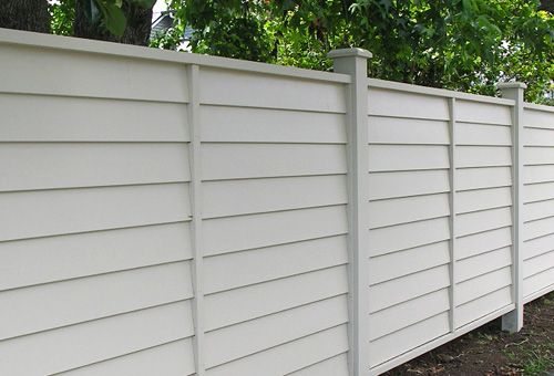 a long white fence is next to some trees and bushes on the side of a house