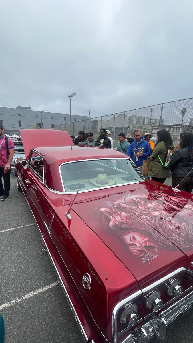 an old red car parked in a parking lot with other cars and people standing around