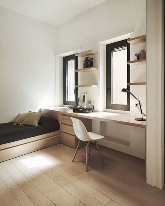 a bedroom with a bed, desk and shelves on the wall next to two windows