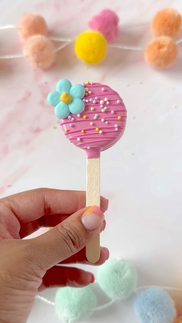 a hand holding a popsicle with sprinkles and flowers on it in front of some pom poms