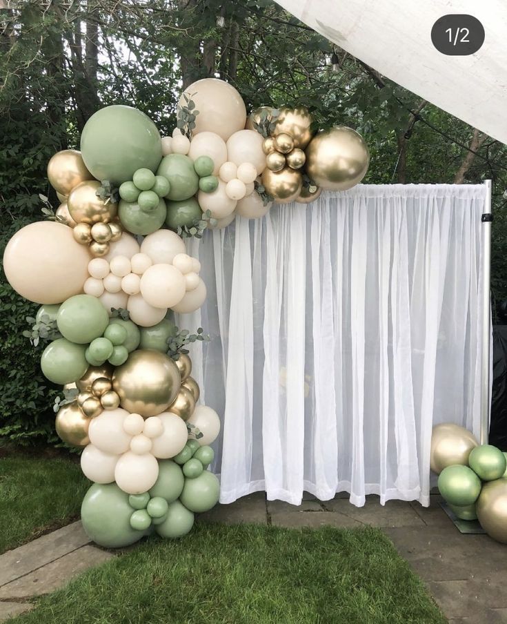 the balloon arch is decorated with gold and green balloons, greenery and white drapes