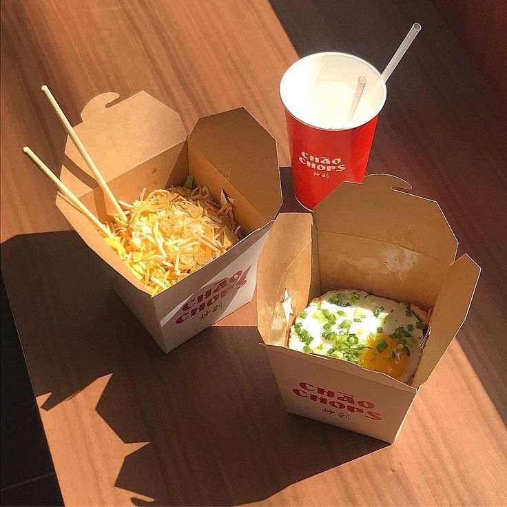 two takeout boxes filled with food sitting on top of a wooden table next to drinks