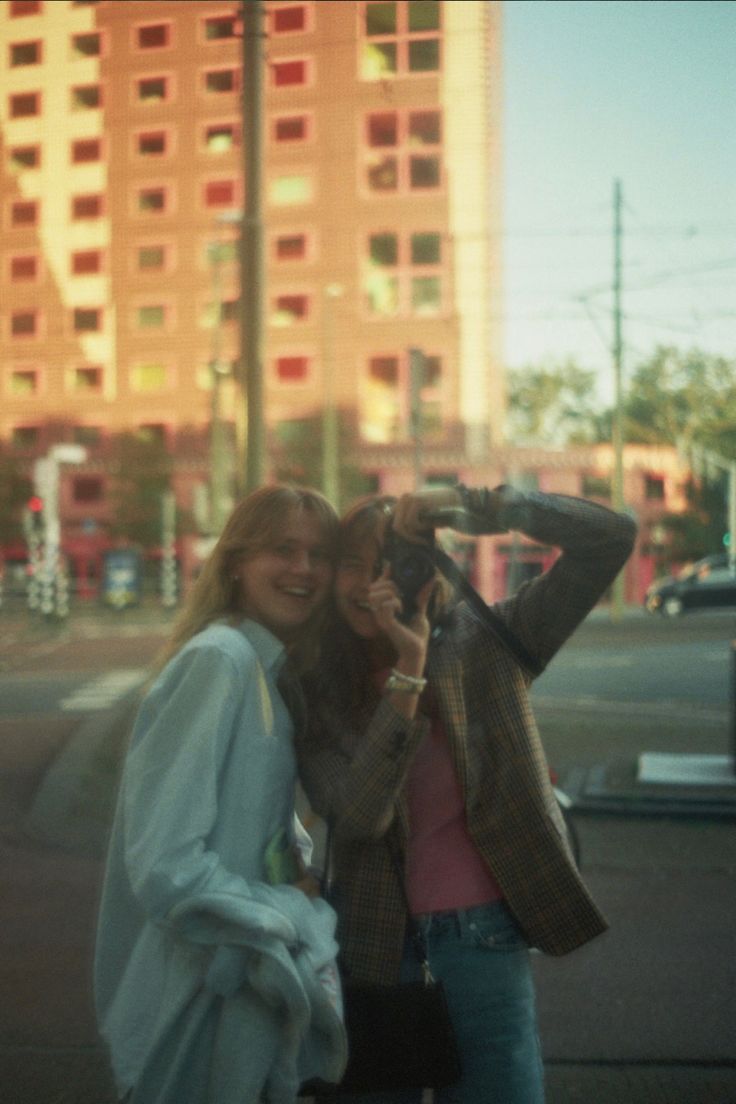 two women standing next to each other in front of a tall building with buildings behind them